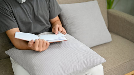 A smart young Asian man reading a book or textbook while relaxing on a comfortable couch in his...