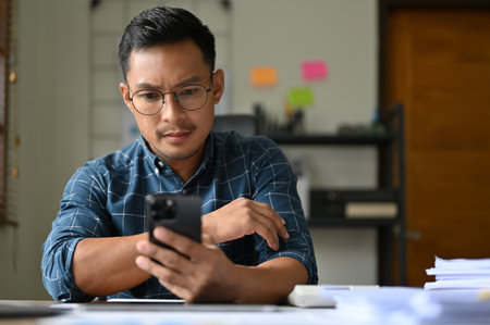 Serious and thoughtful adult Asian businessman or male boss is looking at his smartphone screen, reading a serious email, having an internet connection problem on his phone.の写真素材