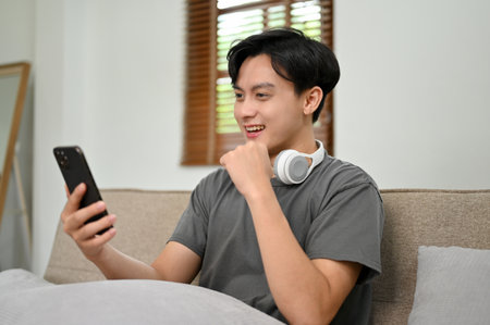 Handsome and happy young Asian man using his smartphone while relaxing on a comfy sofa in his living room. reading an online blog, social media, chatting with friends, online shoppingの写真素材