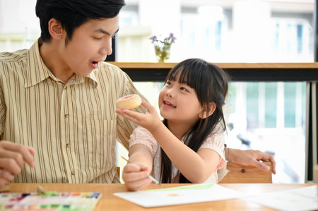 A cute and kind little Asian girl shares a doughnut with her dad in the cafe. enjoying activity, happy family time.の写真素材