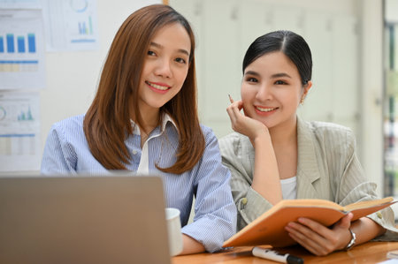 Portrait, Two attractive and charming millennial Asian businesswomen or female office workers sits at office desk, smiling and looking at the camera.の写真素材