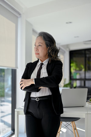 Portrait, Professional and successful Asian aged businesswoman or female CEO in formal business suit arms crosses, looking out the window, daydreaming and pondering, standing in her office.の写真素材
