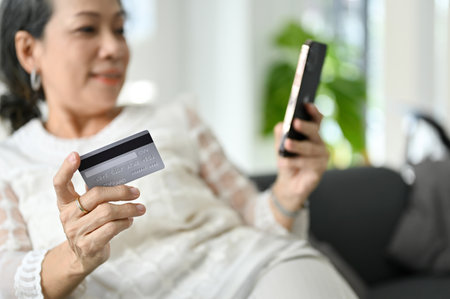selective focus, A beautiful Asian aged woman holding a credit card while relaxing on her sofa and using her smartphone to shopping online.の写真素材