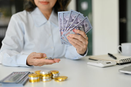 A successful and wealth Asian aged female holding US dollar bills and coins, stack of golden and silver coins and a wad of US dollar bills on the table. cropped imageの写真素材