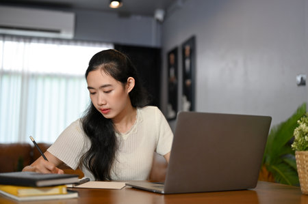 Smart young Asian female college student concentrating doing her homework in her home living room, writing the answer in her school notebook and researching some details on the internet on her laptop.の写真素材