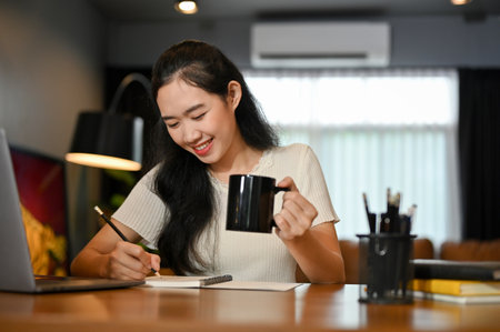 Cheerful young Asian female college student or freelancer working in the living room, sipping morning coffee while writing something on her notebook.の写真素材