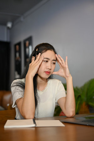 Portrait, Stressed and serious young Asian female suffering from headache, worried about her project, pensively thinking the plan while using a laptop computer.の写真素材