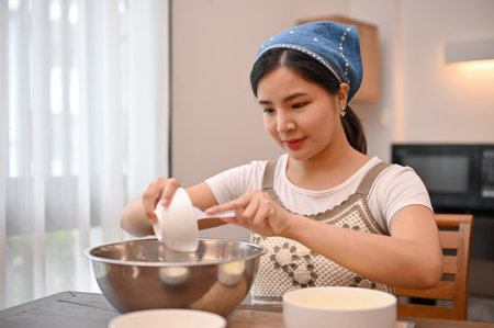 Attractive young Asian female makes a homemade cupcake in her home kitchen, adding sugar in the mixing bowl. cupcake recipe conceptの写真素材