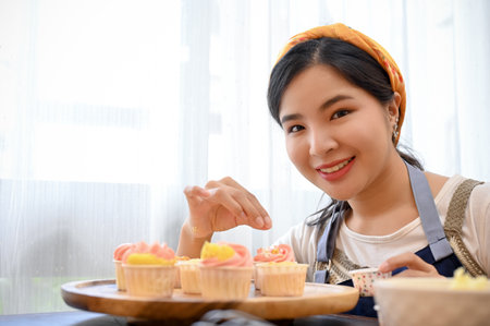 Beautiful young Asian female baker decorating her beautiful homemade cupcake with tiny white chocolate ball.の写真素材