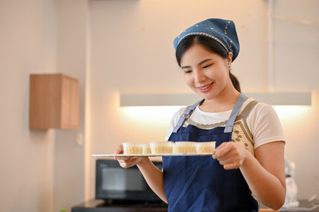Beautiful charming young Asian female baker holding a tray of raw homemade cupcake, preparing to bake in the oven.の写真素材