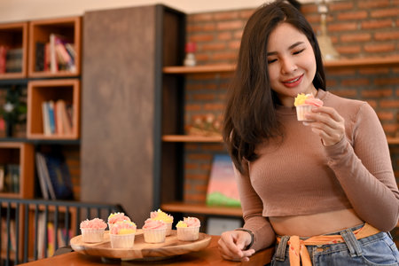 Young beautiful Asian woman eating cupcakes in living room, full of joy and happyの写真素材