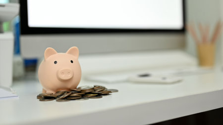 Piggy bank and coins on office desk with blurred background of computer and office supplies....