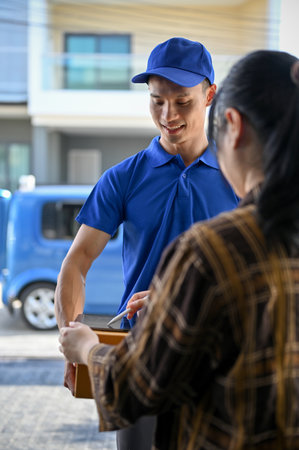 Young Asian delivery man wearing blue uniform handing parcel to customer and have customer sign on tabletの写真素材