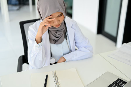 Stressed Asian Muslim female doctor concentrating on her medical research, working at her desk in the doctor's office.の写真素材