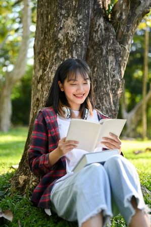 Portrait, Pretty and happy young Asian female college student reading a book while relaxes sitting under the tree in the park.の写真素材