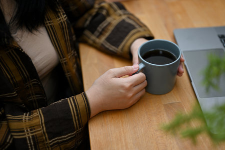 cropped shot, A hipster Asian woman in flannel shirt holding coffee mug, sitting at her office desk.