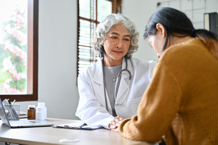 Professional and caring Asian aged female doctor talking, having a serious conversation with her young patient in the hospital office.の写真素材