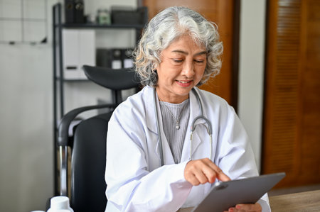 Happy and professional Asian aged female doctor checking her schedule, reviewing medical record on her digital tablet.の写真素材
