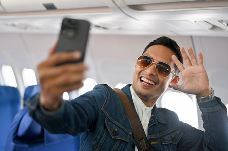 Handsome and joyful Asian male travel blogger or tourist wearing sunglasses taking selfie and recording video of himself by his smartphone during the flight. vacation, holiday, transportationの写真素材