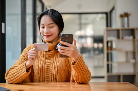 Beautiful young Asian woman sits at her table using her smartphone and credit card to pay an online stuff.の写真素材