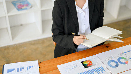 Professional female accountant or financial analyst taking notes on her book, analyzing financial data in her office. cropped, top view.の写真素材