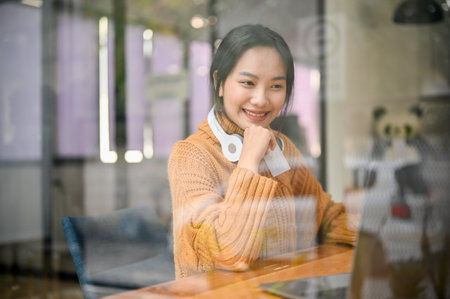Attractive and charming young Asian woman with wireless headphones looking at laptop screen, sitting by the window in the coffee shop. remote working, freelancer, urban lifestyleの写真素材