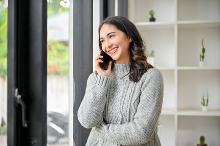 Beautiful and happy Asian woman in cozy sweater stands by the window, looking outside, daydreaming, in-love, talking on the phone with her boyfriend.の写真素材