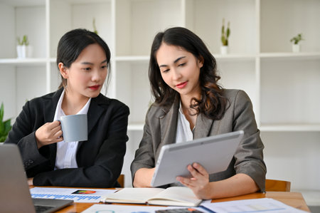 Two professional millennial Asian businesswomen working, looking at tablet screen, discussing and planning project together in the office.の写真素材