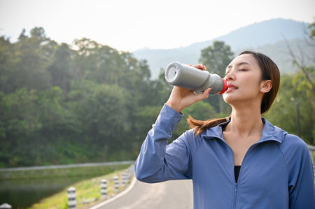 Thirsty and tired Asian woman in sportswear drinking water, resting after running at the beautiful...