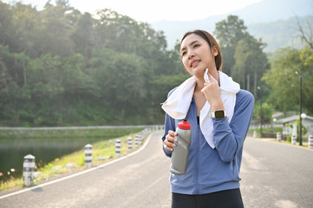 Tired and sweaty Asian woman in sportswear holding a bottle of water, wiping sweat from her face,...