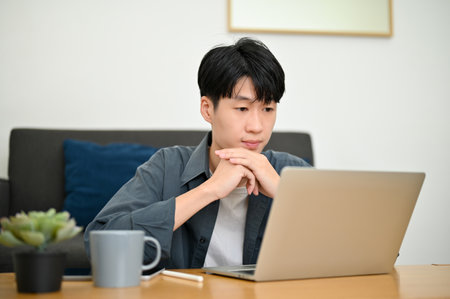 Smart and handsome young Asian male college student or freelancer is in the living room, hands on chin, looking at his laptop screen, thinking about his project.の写真素材