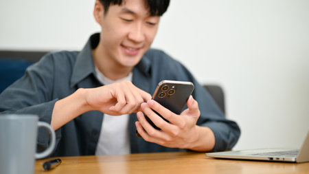Cheerful young Asian man holding a phone, using a mobile application on his smartphone, scrolling on his social media platform, chatting with friends while relaxing in the living room.の写真素材