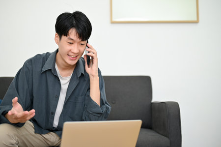 Happy young Asian man looking at laptop screen, talking on the phone with his colleagues or friends, explaining something over the call.の写真素材