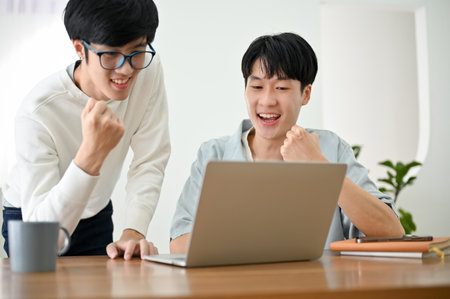Two of young Asian male best friends are showing clenched fist, celebrating success, project winning, receiving good news, winning lottery.の写真素材