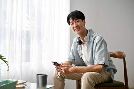 Handsome and attractive young Asian man sits on a chair in minimal living room holding a smartphone, smiling and looking at the camera.の写真素材