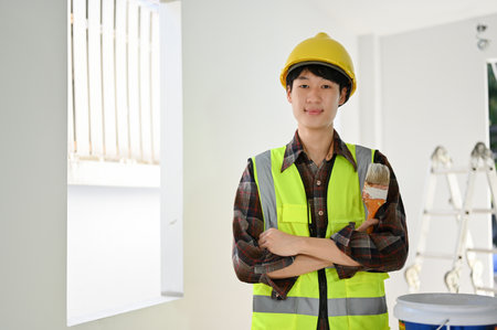 Smart and professional young Asian male construction worker in uniform and yellow hardhat stands with arms crossed, holding a paint brush, smiling and looking at camera.の写真素材