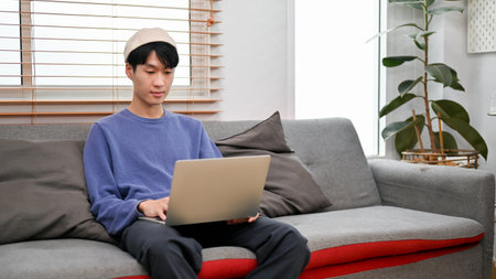 Handsome young Asian man in cosy clothes using laptop computer on sofa in his minimal living room.の写真素材