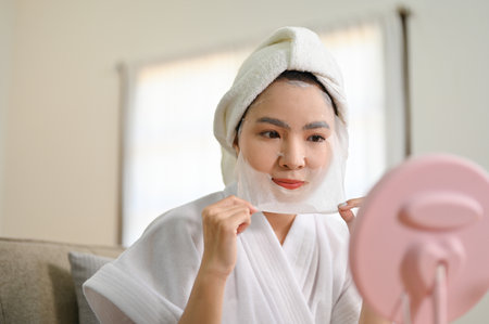 Beautiful young Asian woman looking at the mirror, applying facial sheet mask on her face, having self care day at home. Wellbeing conceptの写真素材
