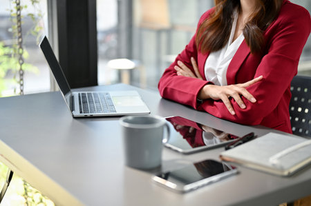 Professional and determined millennial Asian businesswoman or female boss sits at her desk with arms crossed. cropped imageの写真素材