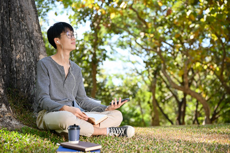 Happy and relaxed young Asian male college student listening to music while relaxing under the tree...