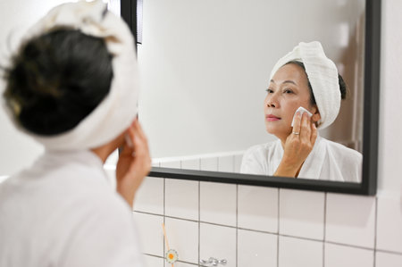 Attractive mature aged Asian woman cleansing face skin with cleanser oil and cotton pad, removing makeup, looking in the mirror. beauty night routine conceptの写真素材