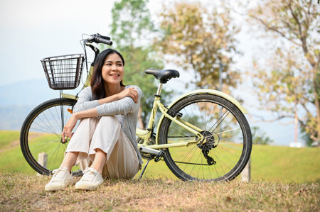 Happy and chill young Asian woman relaxes sitting on grass, taking a rest after cycling at the beautiful nature park. outdoor activity and leisure conceptの写真素材