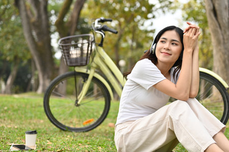 Happy and relaxed young Asian woman in casual comfy clothes listening to music through her wireless headphones and taking a rest after cycling in the public park.の写真素材