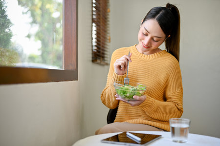 Healthy and beautiful millennial Asian woman in cozy sweater eating delicious and healthy salad bowl at her dining table by the window. healthy lifestyle and wellbeing conceptの写真素材