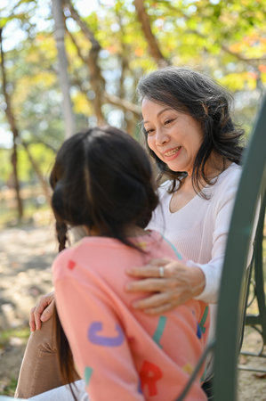 Caring and kind Asian grandmother tells a story to her granddaughter while relax on a bench in the...