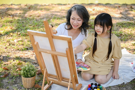 Happy and lovely Asian grandmother teaching her granddaughter to paint watercolors on canvas, having a great time together in the beautiful park.の写真素材