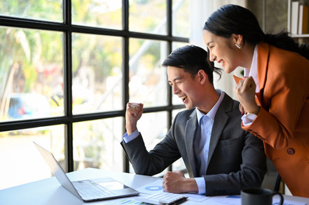 Two happy and cheerful millennial Asian businesspeople showing clenched fist, looking at laptop...