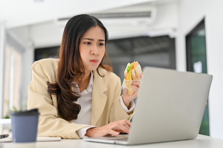 Busy and overworked millennial Asian businesswoman eating a sandwich, having breakfast while managing her project on laptop before deadline. busy business life conceptの写真素材