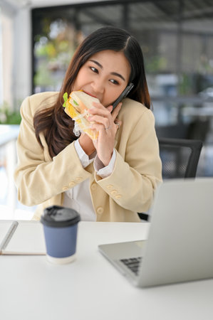 Busy millennial Asian businesswoman eating a sandwich while looking at some details on laptop screen and talking on the phone with her client. multitasking, deadline, hardworking woman, overworkの写真素材