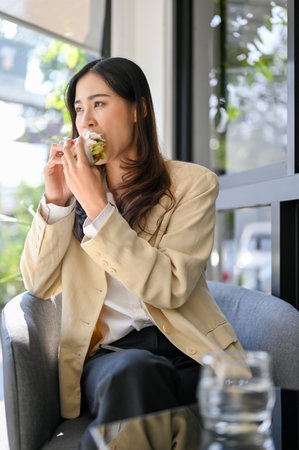 Hungry millennial Asian businesswoman or female office worker having an easy breakfast at the cafe, eating a tasty and healthy sandwich.の写真素材